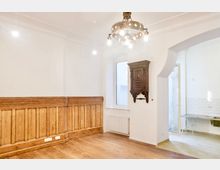 Heller, weitgehend leerer Innenraum mit weißen Wänden, Holzdielenboden und einer langen Holzvertäfelung entlang der linken Wand. An der Decke hängt ein Kronleuchter, neben einem Fenster ist ein dunkler Wandschrank montiert, und ein Rundbogen führt in einen gefliesten Nebenraum mit Glastür.