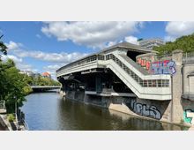 Bahnhof der Hochbahn in städtischer Umgebung, an einem Kanal gelegen. Der Bahnhof hat eine markante erhöhte Struktur mit seitlichen Treppenaufgängen, Graffiti an den Wänden und umliegender Vegetation sowie Brücken im Hintergrund.