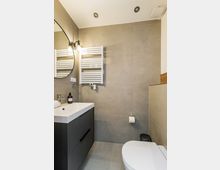 Modernes Badezimmer mit großformatigen grauen Fliesen, Waschtisch mit schwarzem Unterschrank und rundem Wandspiegel mit Leuchten. An der Wand hängen ein weißer Handtuchheizkörper und eine Toilette; Einbaustrahler befinden sich in der Decke.