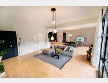 Geräumiger, moderner Wohnraum mit hellen Holzböden, einem grauen Ecksofa auf einem Teppich, einem Glastisch und einem Essbereich im Hintergrund. Große Fenster mit weißen Vorhängen lassen viel Licht herein, und mehrere Hängeleuchten sowie Pflanzen verleihen dem Raum eine wohnliche Atmosphäre.