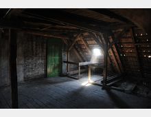 Ein alter Dachboden mit sichtbaren Holzbalken, einer grünen Holztür an einer Ziegelsteinwand und einem kleinen Dachfenster, das Licht hereinlässt. Der Raum hat einen abgenutzten Holzboden und ein teilweise freigelegtes Dachgebälk.