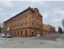 Backstein-Gebäude im industriellen Stil mit mehreren Etagen und großen, symmetrischen Fenstern. Das Gebäude liegt an einer gepflasterten Straße, die von Baustellenabsperrungen und einem geparkten Transporter flankiert ist, unter einem bewölkten Himmel.