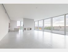 Großzügiger, moderner Innenraum mit weißen Wänden, großen Fensterfronten und einem minimalistisch gestalteten Kamin in der Mitte. Der Raum hat einen grauen, glatten Boden und bietet Ausblick auf eine städtische Umgebung.