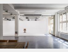 Heller Innenraum eines Fotostudios mit einem weißen Hohlkehlen-Hintergrund und einem zentral platzierten Holzstuhl. An der Decke befinden sich Studioleuchten, und auf der rechten Seite sind große Fenster mit Vorhängen zu sehen, die Tageslicht hereinlassen.