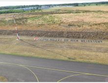 Eine grasbewachsene Fläche neben einer Landebahn mit einem rot-weißen Beleuchtungsmast; am Boden steht in großen Buchstaben: „Lena, willst du mich heiraten?“ mit einem roten Herzsymbol daneben. Im Hintergrund sind Felder und vereinzelte Gebäude zu sehen.