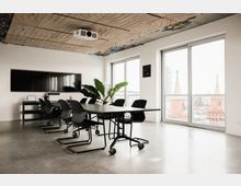 Modernes Besprechungszimmer mit einem großen rechteckigen Tisch und mehreren schwarzen Stühlen auf einem Betonboden. An der Wand hängt ein großer Bildschirm, die Decke hat ein abstraktes Design, und durch große Fenster ist ein Blick auf rote Gebäudespitzen und die Stadt sichtbar.