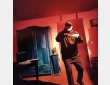 Ein Raum mit rotem Licht, in dem ein Mann in dunkler Kleidung gestikuliert. Im Hintergrund stehen ein blauer Kleiderschrank und ein Tisch mit einem kleinen Gegenstand darauf, die Szene wirkt leicht düster beleuchtet.