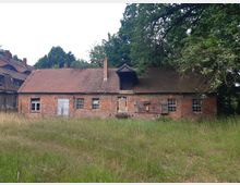 Altes Backsteingebäude mit roten Ziegelsteinen und einem Ziegeldach, teils verfallenen Fenstern und einer Holztür im Vordergrund, umgeben von hohem Gras und Bäumen im Hintergrund. Links im Bild ist ein weiteres Gebäude teilweise sichtbar.