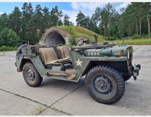 Ein grüner, offener Militär-Jeep mit US-Armee-Markierungen steht auf einer betonierten Fläche. Im Hintergrund ist ein alter, überwucherter Hangarbunker vor einem Wald aus Nadelbäumen zu sehen.