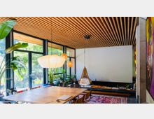 Ein modernes Wohnzimmer mit großen Glasfenstern, die Ausblick auf einen Garten bieten. Holzlamellen an der Decke, ein großer Esstisch, hängende Papierlampen und ein Hängesessel prägen den Raum, der mit Pflanzen und farbigen Teppichen dekoriert ist.