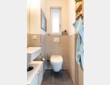 Kleines Badezimmer mit beigefarbenen Wandfliesen und dunklem Bodenfliesen. Zentral befindet sich eine Hänge-Toilette unter einem schmalen Fenster, links ein kleines Waschbecken mit Unterschrank und rechts ein Holzregal und ein Handtuchhaken.