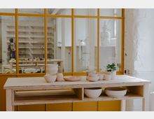 Ein heller Raum mit einem Holztisch, auf dem verschiedene handgefertigte Keramikgefäße und eine kleine Zimmerpflanze stehen. Im Hintergrund befindet sich eine gelb umrahmte Glaswand, durch die Regale und weitere Gegenstände sichtbar sind.