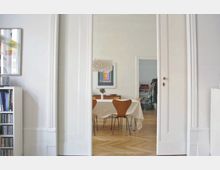 Innenraum einer Wohnung mit hellem Holzboden und weißen Wänden, die durch eine große Türöffnung verbunden sind. Im hinteren Raum steht ein Esstisch mit heller Tischdecke, umgeben von Holzstühlen, sowie eine originelle Hängelampe und ein buntes Wandbild über einer Kommode.