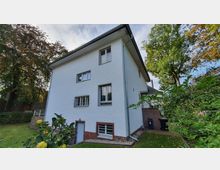 Zweistöckiges Einfamilienhaus mit weiß verputzter Fassade und gemauertem Sockelbereich aus roten Ziegelsteinen. Umgeben von einem Garten mit Obstbäumen und Rasenfläche, im Hintergrund hohe Laubbäume.