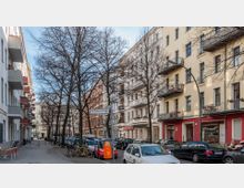 Straßenansicht einer urbanen Wohngegend mit mehrstöckigen Altbauhäusern, die Balkone und große Fenster besitzen; Bäume stehen entlang des Gehwegs, und geparkte Autos sowie Fahrräder säumen die Straße. Ein Straßenschild mit der Aufschrift "Kyffhäuserstraße" ist sichtbar.