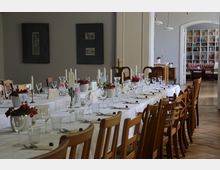 Ein langer, festlich gedeckter Tisch in einem hellen Raum mit Holzfußboden, weißen Tischdecken, Kerzenständern, Blumendekorationen und Gläsern. Im Hintergrund sind ein Bücherregal, ein Sofa und durch einen Türbogen weitere Sitzgelegenheiten zu sehen.