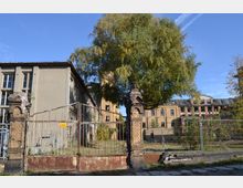 Ehemaliges Industriegelände mit verfallenen Backsteingebäuden, einem rostigen Tor und einem teils zerstörten Mauerpfeiler. Im Hintergrund stehen unterschiedliche Gebäude, teils mit eingefallenen Fenstern, in einem verwilderten Umfeld.