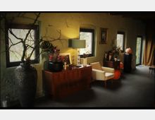 Wohnraum mit rustikalem Stil, grün gestrichenen Wänden und mehreren Fenstern, die natürliches Licht hereinlassen. Der Raum ist dekoriert mit großen Vasen, einer Kommode mit Lampe und Blumenarrangements, sowie einem Sessel und herbstlichen Akzenten wie Kürbissen.