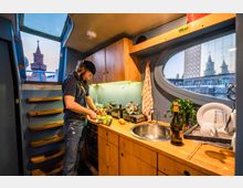 Ein Mann arbeitet in einer kleinen Küche mit Holzschränken und ovalem Fenster in einem Hausboot. Durch das Fenster sind der Fluss, Gebäude und ein Turm zu sehen.