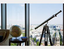 Ein Innenraum mit einem runden Tisch, auf dem ein Globus und ein Buch stehen, neben einem Teleskop auf einem Stativ. Durch die großen Fenster ist eine Stadtlandschaft mit modernen Gebäuden und einer Kirche mit grünem Dach zu sehen.
