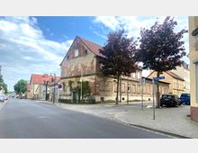 Straßenszene in einer Kleinstadt mit einem alten, verfallenen Gebäude aus Backstein an einer Straßenecke, umgeben von gepflasterten Gehwegen und Bäumen. Im Hintergrund befinden sich weitere Wohnhäuser mit roten Ziegeldächern, Straßenschilder und geparkte Autos.