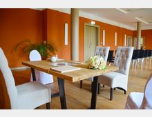 Ein festlich dekorierter Raum mit orangefarbenen Wänden und hellem Holzboden, ausgestattet mit einem Holztisch und gepolsterten Stühlen. Auf dem Tisch liegen ein Blumenarrangement und Dokumente; im Hintergrund sind weitere Stühle sowie eine Tür zu sehen.