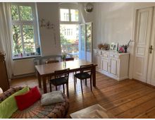 Wohnzimmer mit Holzboden, großem Holztisch und Stühlen, einer bunt gemusterten Couch mit Kissen und einer weißen Kommode. Große Fenster und eine offene Balkontür lassen Tageslicht herein, mit Blick auf Bäume und ein gegenüberliegendes Gebäude.