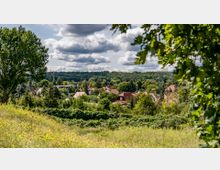 Blick auf eine ländliche Siedlung mit Häusern, roten Dächern und vielen Bäumen, umgeben von grünen Wiesen und dichter Vegetation; im Hintergrund erstreckt sich ein bewaldetes Gebiet unter einem teilweise bewölkten Himmel.