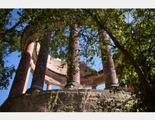 Rundbau aus rotem Backstein mit mehreren Säulen auf einem steinernen Sockel, teilweise verdeckt durch umliegende Bäume und Laub; blauer Himmel im Hintergrund.