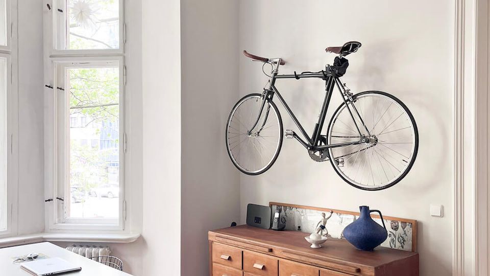 Bright, minimalist living room with high ceilings and ornate plaster molding, featuring tall white-framed windows and light wood floors. A wall-mounted bicycle hangs above a long wooden sideboard, with a white desk and chairs near the window and a sofa edge in the foreground.