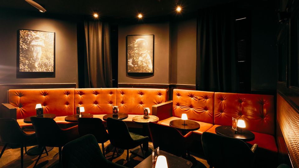 Dunkler Lounge- oder Barbereich mit langen, orangefarbenen, gepolsterten Eckbänken, kleinen runden Tischen mit Tischlampen und schwarzen Stühlen. An den Wänden hängen gerahmte Bilder, und Vorhänge sowie Spotbeleuchtung prägen die gedämpfte Atmosphäre.