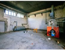 Ein leerer, industrieller Raum mit Betonboden und Wänden, einer schwarzen Wellblechdecke sowie großen Fenstern nahe der Decke. Im Raum befinden sich eine rote Heizungsanlage mit Rohren und ein liegender Metalltank an der Wand.