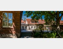 Innenhof mit gepflasterter Terrasse und kleiner Treppe zwischen einem Backsteingebäude mit großen Sprossenfenstern und einem hellen Gebäude mit roten Ziegeldächern. Rechts steht ein großer Baum mit Grünfläche und Hecken; im Hintergrund ragt ein Kirchturm über die Dächer.