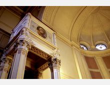 Innenansicht eines historischen Kirchenraums mit verzierten Säulen und einer Kanzel, die durch Reliefs und ein Porträt geschmückt ist. Im Hintergrund ist eine reich verzierte Kuppeldecke mit Rundfenstern und farbigem Glas zu sehen.