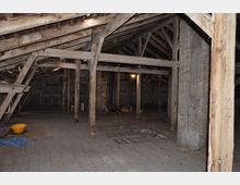 Dachboden eines alten Gebäudes mit sichtbarer Holzkonstruktion, schrägen Balken und teilweise beschädigten Wänden. Der Raum ist spärlich beleuchtet, enthält verstreute Gegenstände wie einen gelben Eimer und blaue Folie, und der Boden besteht aus abgenutzten Holzdielen.