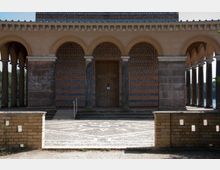 Fassade eines historischen Gebäudes mit einer zentralen Holztür, flankiert von Bögen mit steinernen Säulen und dekorativen, gemusterten Ziegeln in Blau- und Ockertönen. Im Vordergrund ein gepflasterter Platz mit geometrischen Mustern und eine niedrige Backsteinmauer.