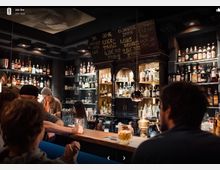 Eine stimmungsvolle Bar mit dunkler Holzeinrichtung, einer gut gefüllten Rückwand mit Spirituosenflaschen und Chalkboard-Menüs. Im Vordergrund sitzen Gäste an einer Theke, hinter der Mitarbeitende Drinks zubereiten.