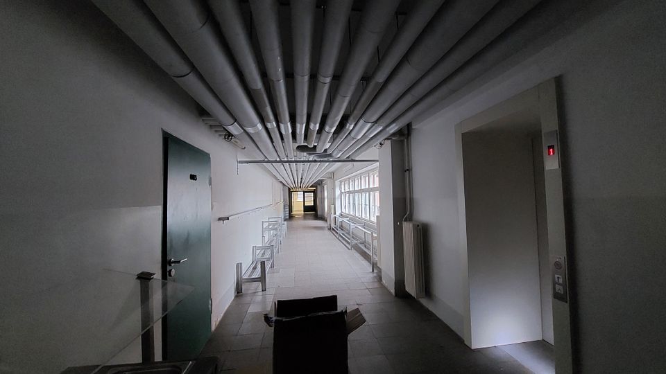 Dunkler Innenflur mit freiliegenden Rohrleitungen an der Decke, einer geschlossenen grünen Tür auf der linken Seite und einem Aufzug mit einer beleuchteten "1" rechts. Auf der rechten Wandseite befinden sich Fenster und Heizkörper, die Licht in den Raum lassen.