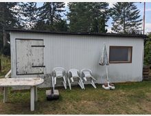 Kleines, einstöckiges Holzhaus mit flachem Dach und hellgrauer Fassade in einem Garten, umgeben von hohen Bäumen. Vor der Wand stehen gestapelte weiße Plastikstühle und ein geschlossener Sonnenschirm; links ist eine verriegelte Tür, rechts ein Fenster.