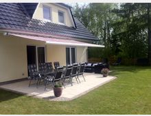 Ein Einfamilienhaus mit gelber Fassade und dunklem Dach, das eine Terrasse mit gepflastertem Boden zeigt. Auf der Terrasse stehen ein großer Tisch mit mehreren Stühlen sowie ein Sonnenschutz, während der angrenzende Garten von Rasen und Bäumen umgeben ist.