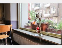 Ein schlicht eingerichtetes Café mit einem Holztisch und Stuhl in der linken Ecke. Auf der Fensterbank steht eine weiße Tischlampe mit einer roten und einer gedimmten Glühbirne, neben Blumenkästen vor einem großen Fenster mit Blick auf eine Altbaufassade.