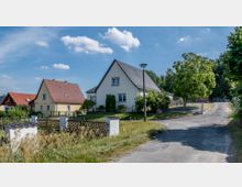 Wohnsiedlung mit freistehenden Einfamilienhäusern, erkennbar an den Satteldächern und hellen Fassaden. Im Vordergrund ein Garten mit Holzzaun, daneben eine asphaltierte Straße, umgeben von Grünflächen und Bäumen.