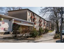 Wohnstraße mit dreistöckigen Wohnblöcken, die Balkone mit roten Geländern haben. Im Vordergrund befindet sich ein kleiner Laden mit Markise, daneben ein Bürgersteig mit Bäumen und parkenden Autos.