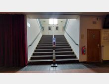 Eine breite Treppe mit dunklen Stufen und beidseitigen Handläufen führt zu einer oberen Etage. An der Wand hängen Porträtbilder, darüber befindet sich ein Fenster, das Tageslicht einlässt. Im Vordergrund steht ein Desinfektionsspender, links befindet sich ein roter Vorhang, rechts eine Tür und ein an der Wand befestigter Feuerlöscher.