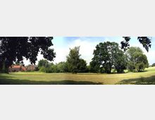 Panorama einer ländlichen Landschaft mit einer Wiese im Vordergrund, Bäumen am Rand und einem roten Ziegeldach eines Bauernhauses links im Bild. Der Himmel ist klar und leicht bewölkt, und der Bereich liegt teilweise im Schatten von Bäumen.