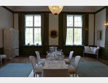 Eleganter Speisesaal mit hohen Fenstern, schweren grünen Vorhängen und verzierten Wänden. Im Zentrum steht ein gedeckter Tisch mit weißen Stühlen, darüber hängt ein Kronleuchter.