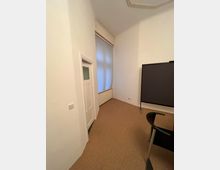 Innenaufnahme eines kleinen Zimmers mit weiß gestrichenen Wänden, einem hohen Fenster mit blickdichtem Rollo und beigefarbenem Teppichboden. Links befindet sich eine Tür, rechts ein Teil eines schwarzen Stuhls und eines Tisches.