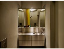 Modernes Badezimmer mit wandhängendem Waschtisch aus Edelstahl und rundem Waschbecken, davor eine große Spiegelwand. Im Hintergrund sind eine Dusche mit gelbem Duschvorhang, eingelassene Nischen und eine Deckenleuchte zu sehen.