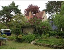 Garten mit viel Grün und Bäumen, ein Haus mit Satteldach und einer Fassade aus hellem Putz im Hintergrund. Ein blühender Fliederbusch, ein blauer Wohnwagen und ein Trampolin sind im Vordergrund zu sehen.