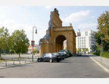 Eine historische Backsteinstruktur mit einem großen Torbogen und dekorativen Verzierungen, umgeben von geparkten Autos und moderner Architektur im Hintergrund. Auf der linken Seite stehen Bäume und eine freie Betonfläche, rechts eine gepflasterte Straße mit Laternen.